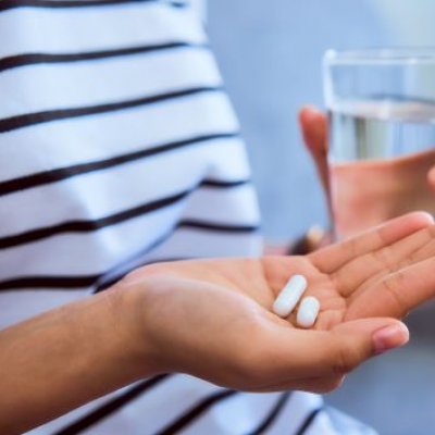 close up of a person's hand holding two small white capsules in the background is their other hand holding a glass of water
